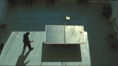 Overhead view of a lone figure holding a paddle beside a worn ping-pong table in a vast, sun-raked hall, long shadows thrown across the concrete floor and the far side of the table empty — a rally waiting for its return.