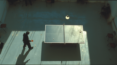 Overhead view of a lone figure holding a paddle beside a worn ping-pong table in a vast, sun-raked hall, long shadows thrown across the concrete floor and the far side of the table empty — a rally waiting for its return.