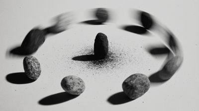 Low-angle black and white photograph of stones in circular formation, outer stones heavily motion-blurred in sweeping arcs while a single sharp stone stands upright at center surrounded by scattered dark particles.