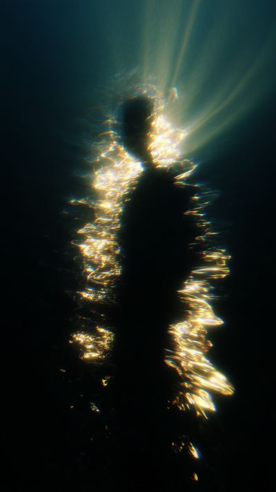 A dark silhouette stands centered in deep blue-black void, defined only by golden light rays streaming from above that fragment and ripple around the figure's edges like underwater caustics.