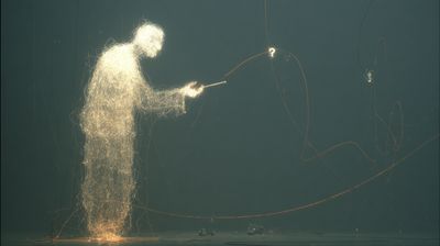 A figure woven from glowing gold filaments stands in profile and extends a thin luminous stylus toward a floating question mark at right, the two joined by a single amber arc. A teal-grey studio, sparks pooling at its feet.