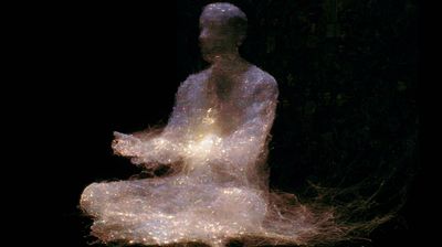 The same seated figure spun from fine warm filaments — gold and pale-white threads woven into a translucent body against pure black, a soft golden light glowing at its centre, faint faces dissolving in the dark behind.