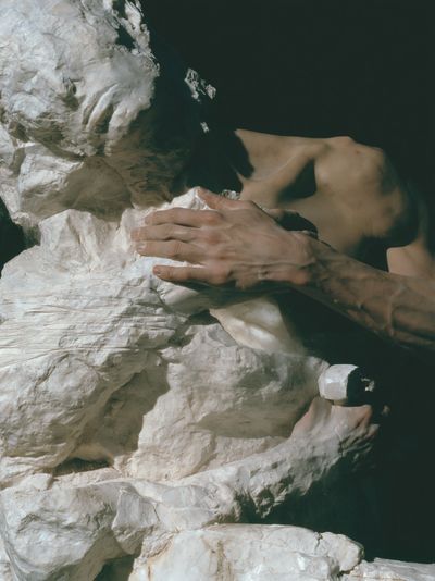 Close view of a veined human hand resting flat on rough white marble, a second hand below gripping a pale chisel.