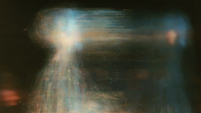 A blurred glowing figure in blue and cream stands in a dark, grainy frame, with faint text lines and an amber blur at right.